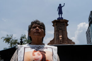 Retrato de defensora María del Carmen Volante Velázquez, con un delantal blanco que lleva impresa la ficha de búsqueda de su hija Guadalupe Pamela Gallardo Volante. De fondo se ve la Glorieta de las mujeres que luchan en Ciudad de México.